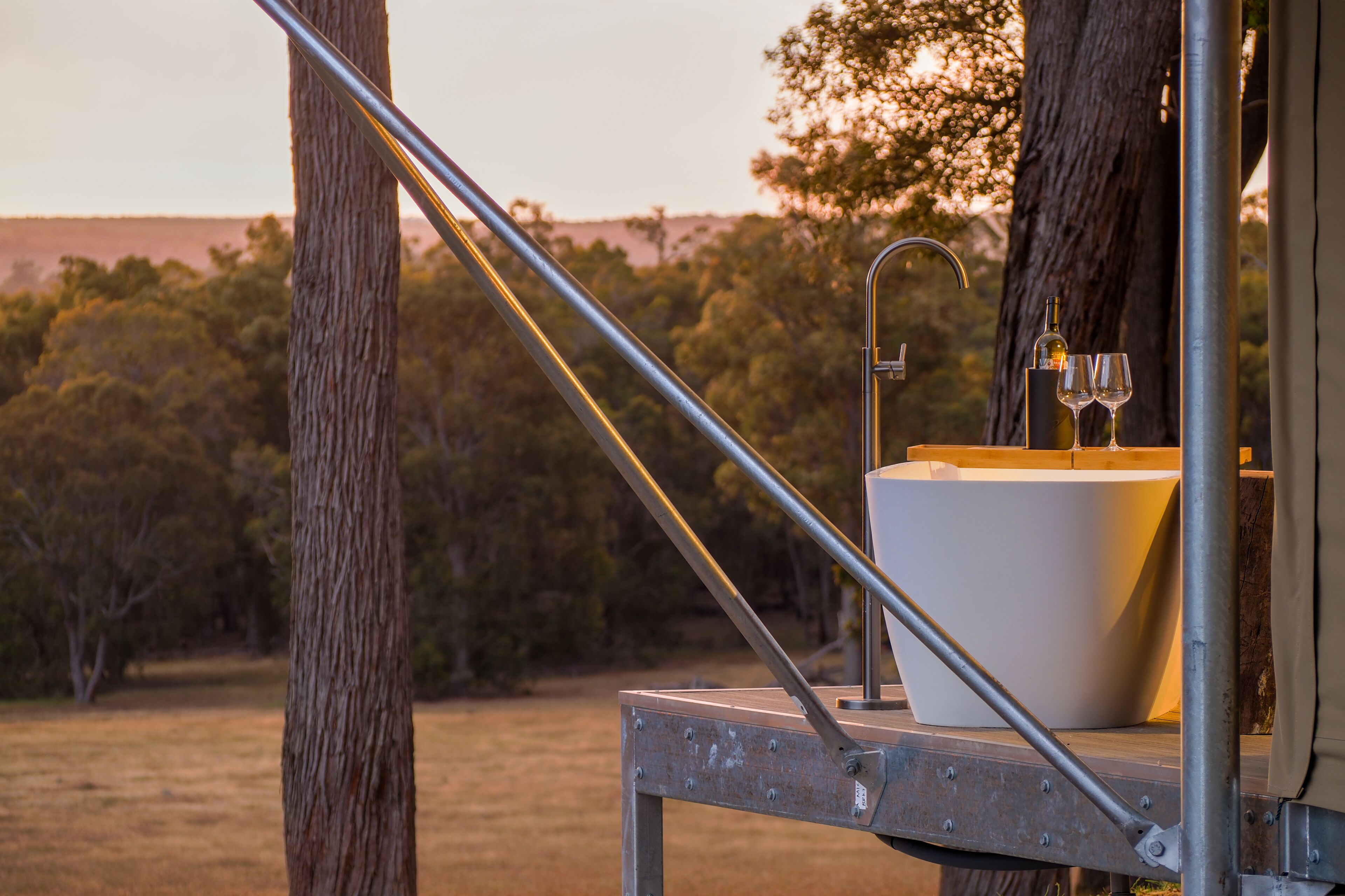 Un baño exterior rústico con un lavabo blanco, una botella y dos vasos, con árboles y un atardecer de fondo.