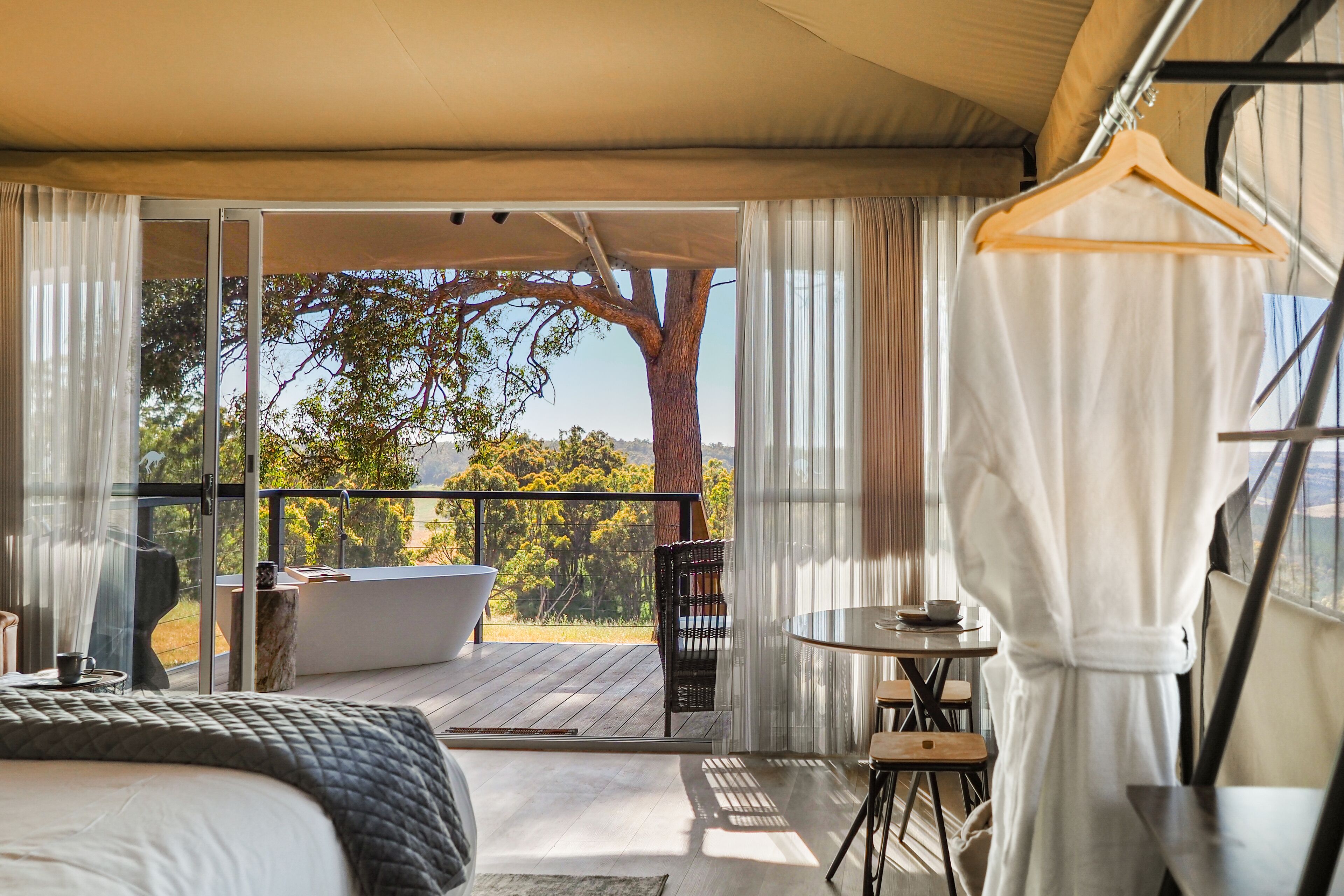 A bedroom with a large bed, a small table, a chair, and a view of a balcony with a tree.