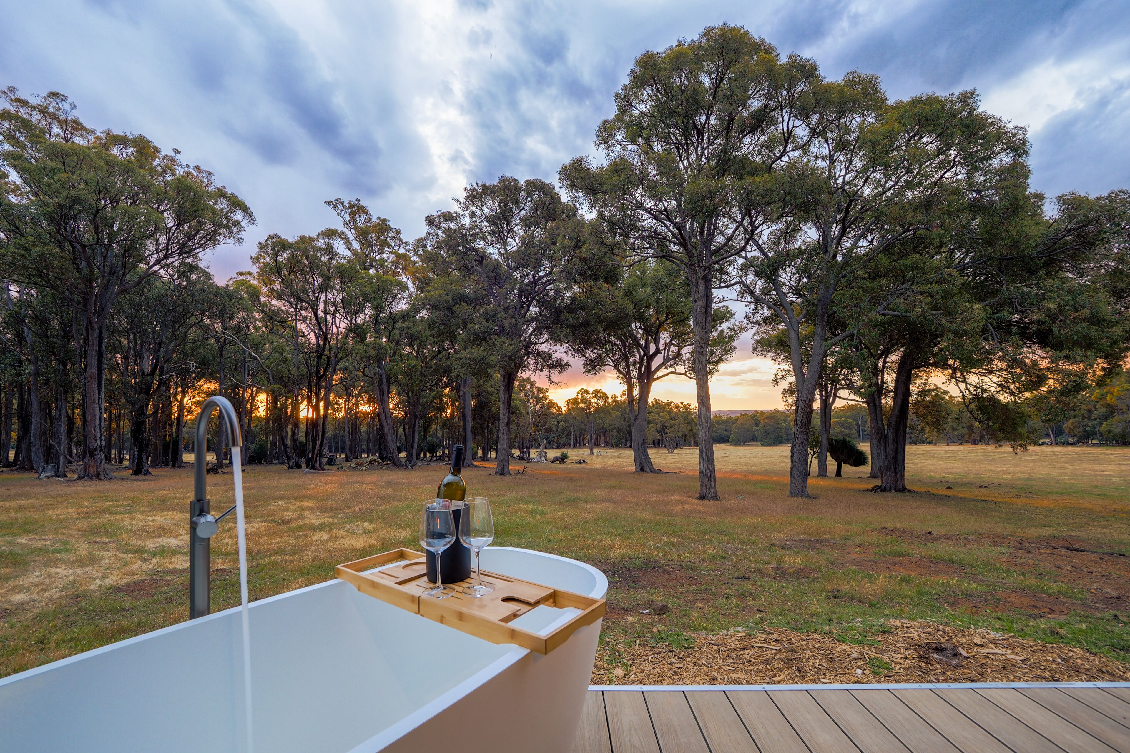A wooden deck with a white bathtub and a faucet, surrounded by trees and a sunset view.