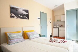 Un dormitorio con una cama, dos almohadas, una mesita de noche, una botella de vino y pétalos de rosa. Cuenta con una zona de cocina con armarios y un lavamanos.