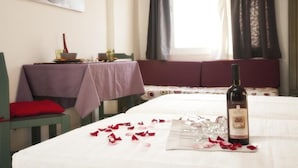 A hotel room with a dining table set with rose petals, a bottle of wine, and glasses. There is a window with curtains, a bed with a red pillow, and a painting on the wall. - Selena View Hotel & Apartments (Hersonissos)