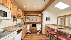 A kitchen with wooden cabinets, a microwave, a stove, and a dining area with stools. - Old Hundred Unit 104 | Sunny, Corner Townhouse, Treetop Views, 3 Blocks to Aspen Mtn (Aspen)