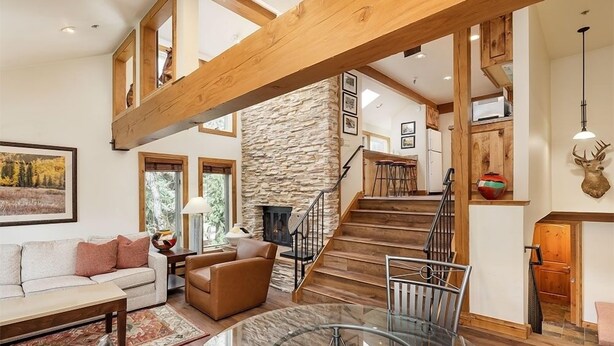 A spacious living area with a stone fireplace, a wooden staircase, and a glass dining table. - Old Hundred Unit 104 | Sunny, Corner Townhouse, Treetop Views, 3 Blocks to Aspen Mtn (Aspen)