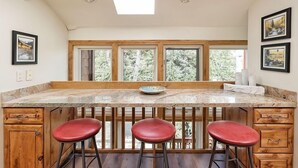 A kitchen with a large island, wooden bar stools, and a view of trees through the windows. - Old Hundred Unit 104 | Sunny, Corner Townhouse, Treetop Views, 3 Blocks to Aspen Mtn (Aspen)