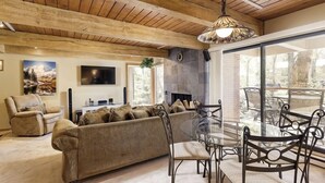 A living room with a sofa, a glass table, and chairs, a fireplace, and a TV on the wall. - Chateau Eau Claire Unit 17 | Serene Condo Overlooking the River with Many Upgrades (Aspen)