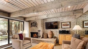 A cozy living room with a fireplace, wooden furniture, and a large window overlooking a garden. - Chateau Eau Claire Unit 8 | Riverfront Condo, Walking Distance to Town (Aspen)