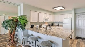A modern kitchen with white cabinets, a granite countertop, a built-in microwave, and a refrigerator. There are three bar stools under the island. - High-Rise Destin Condo w/ Balcony + Pool View (Destin)