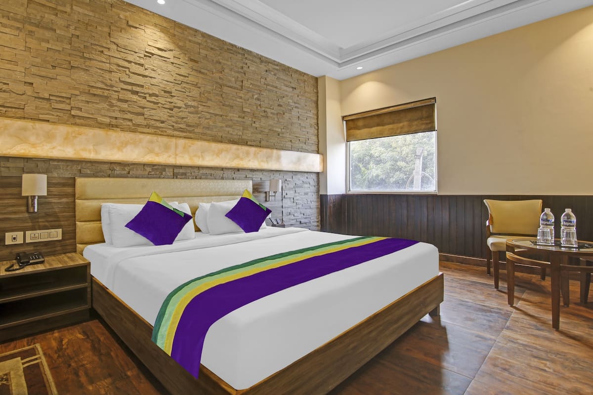 Una habitación de hotel con una cama grande, una mesita de noche, una lámpara, un ventanal con vistas a árboles y una silla.