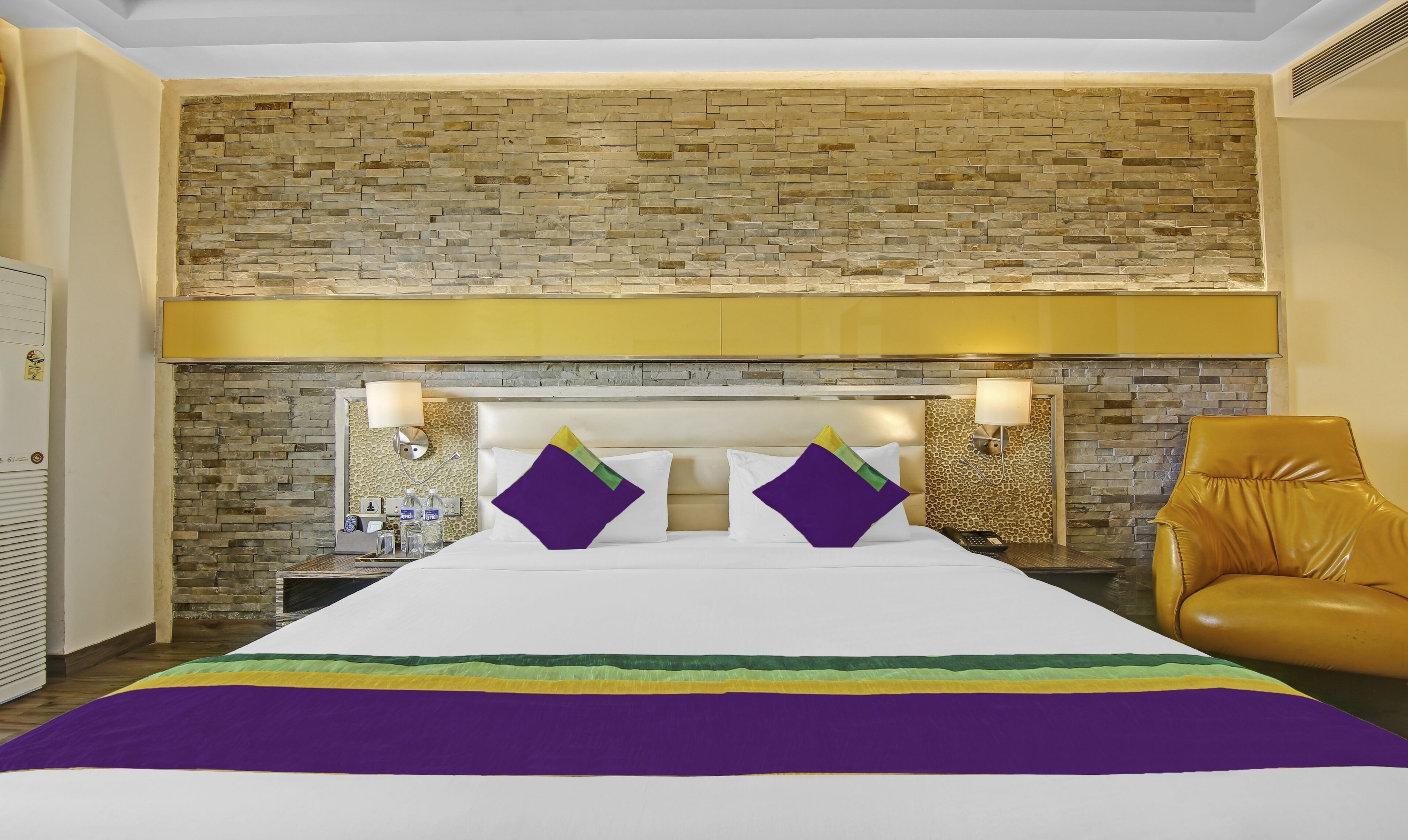 Une chambre d’hôtel avec un mur en pierre, un lit recouvert d’une courtepointe blanche et violette, deux tables de chevet avec des lampes et un fauteuil en cuir.
