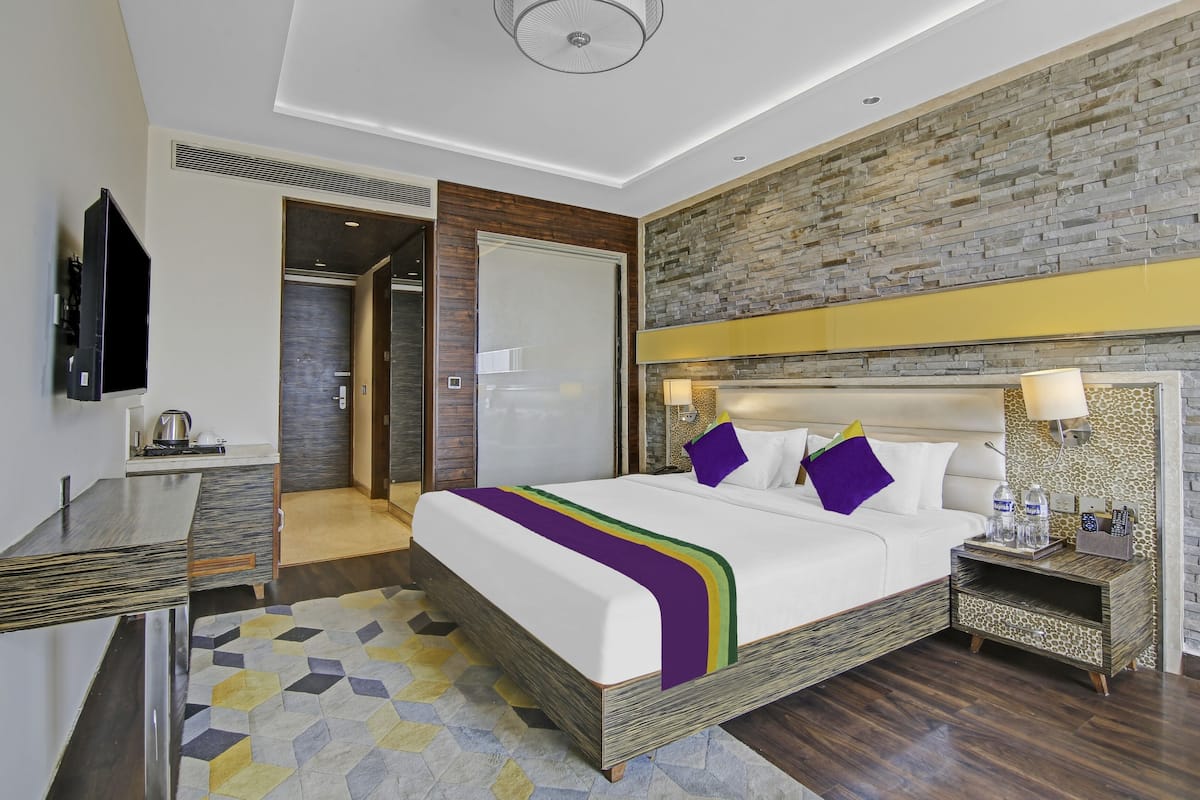 Una habitación de hotel moderna con una cama grande, un televisor de pantalla plana, un escritorio con hervidor y una pared con textura de piedra.