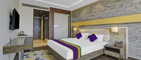 Una habitación de hotel moderna con una cama grande, un televisor de pantalla plana, un escritorio con hervidor y una pared con textura de piedra.