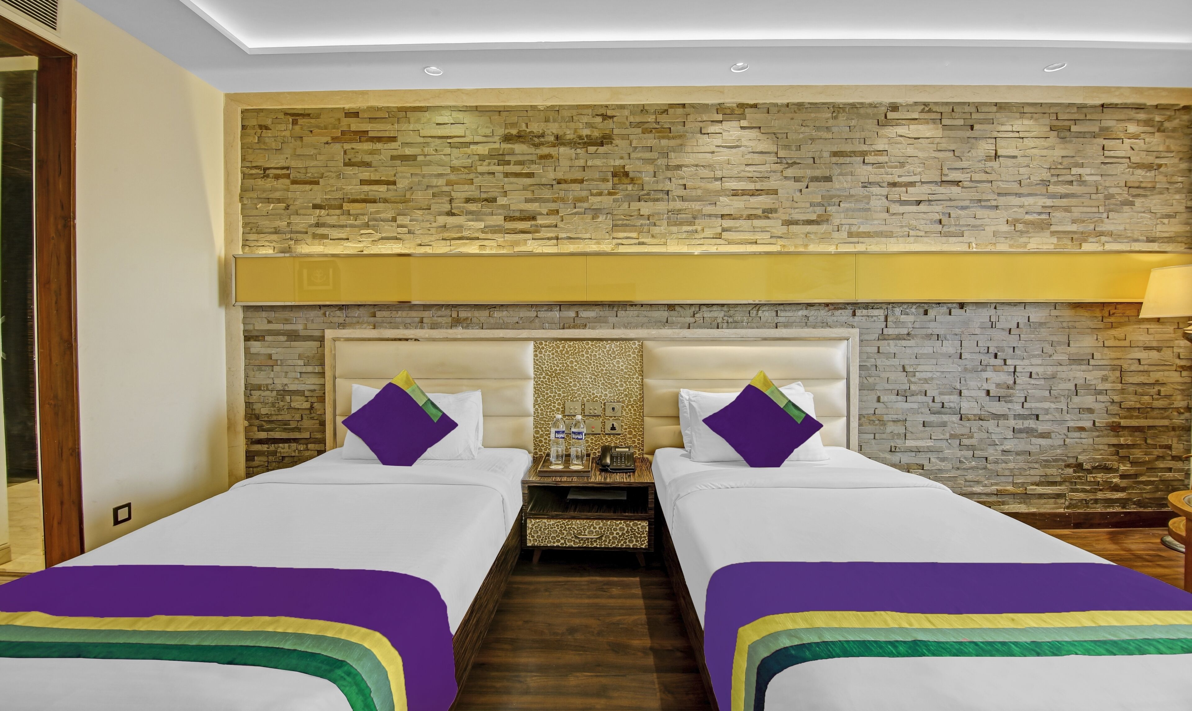 Habitación de hotel con dos camas, pared de piedra, cabecero de madera y mesita de noche con lámpara.