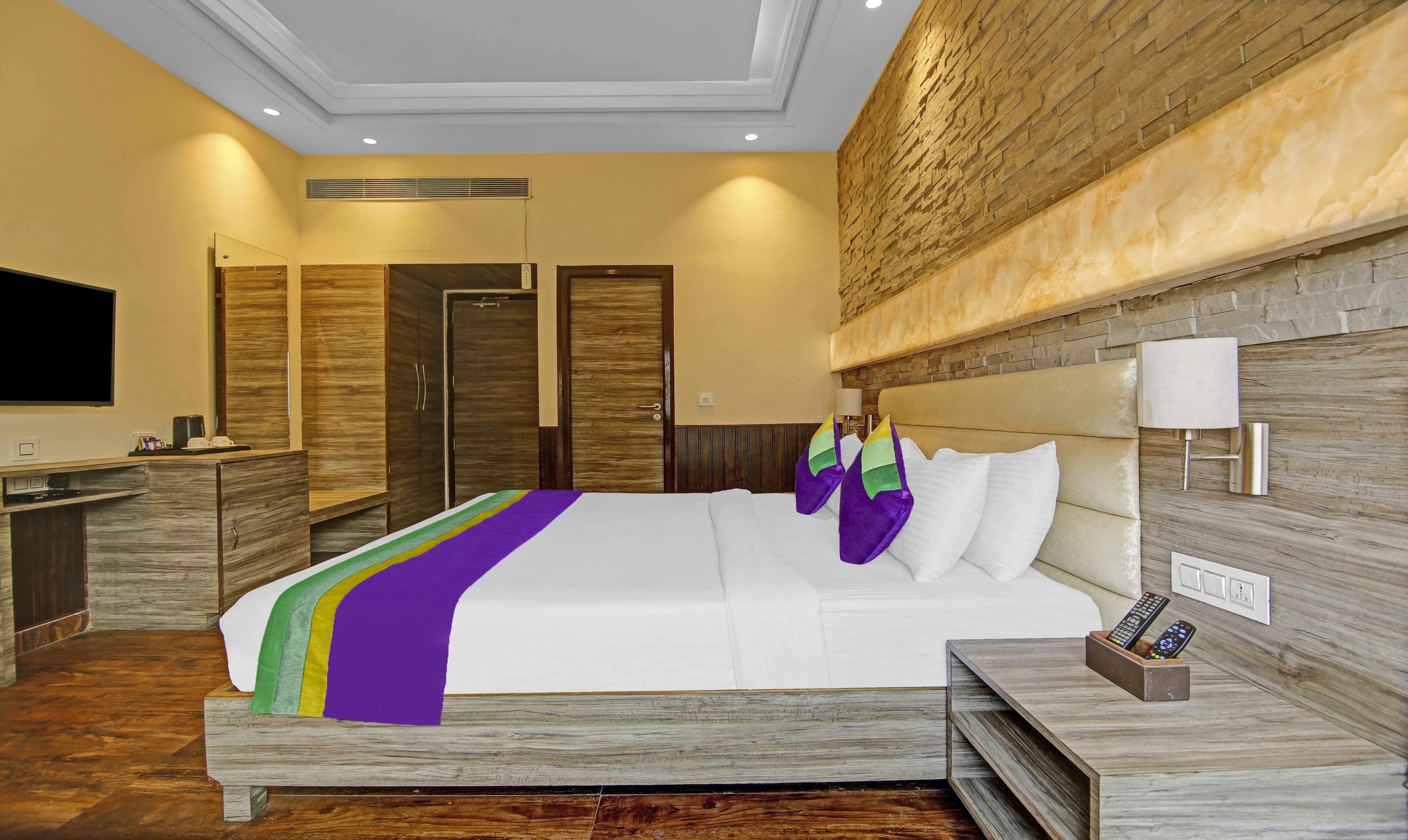 Una habitación de hotel con una cama grande, cabecera de madera, televisor, mesita de noche y lámpara.