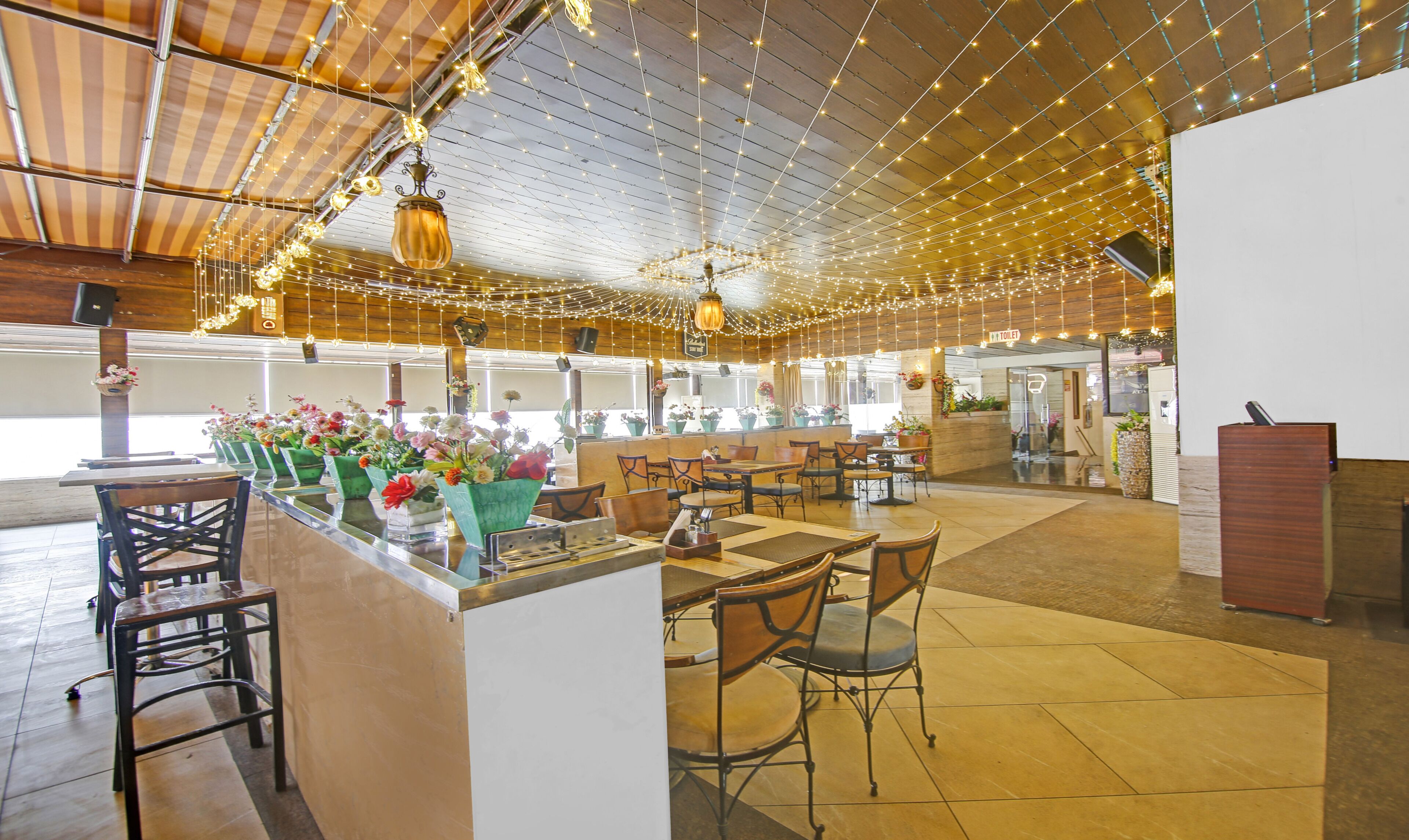 A restaurant with a bar area, multiple tables and chairs, hanging lights, and a view of the kitchen.