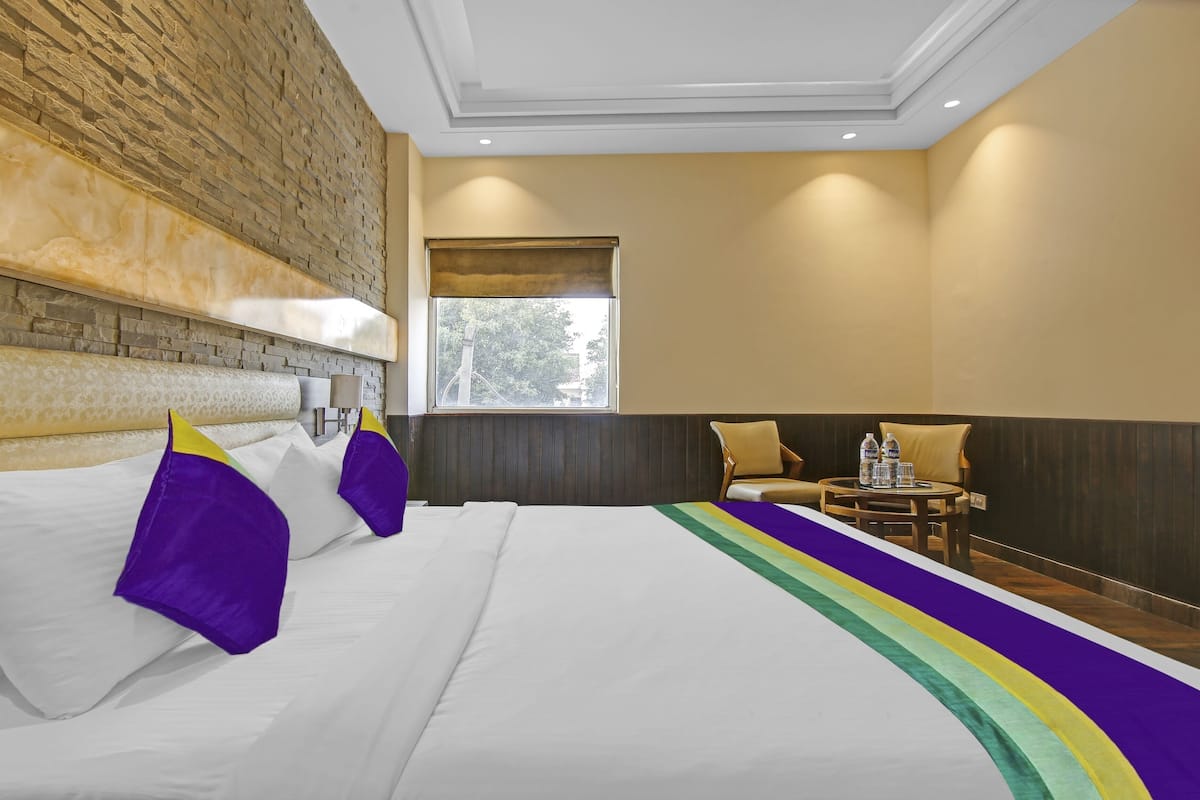 Habitación de hotel con pared de ladrillo decorativa, cama grande con almohadas blancas y púrpuras, mesita con botellas de agua y ventana con vista a árboles.