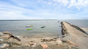 Escena costera con un paseo de concreto que lleva al agua, barcos y una costa distante.