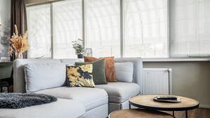 A modern living room with a grey sectional sofa, two round coffee tables, and a window with blinds. - The Lion King (Groningen)