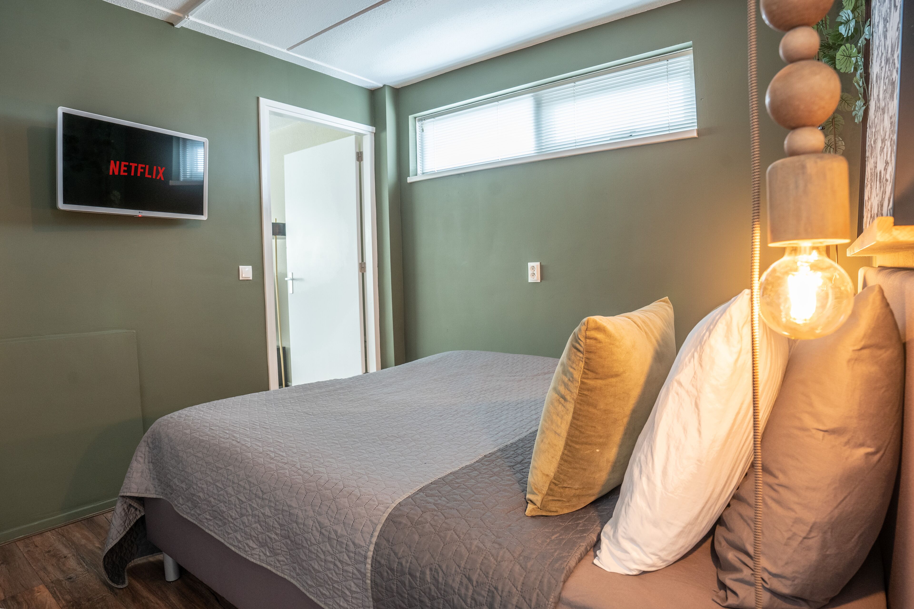 A bedroom with a bed, a television displaying the Netflix logo, a window with blinds, and a wall-mounted lamp.