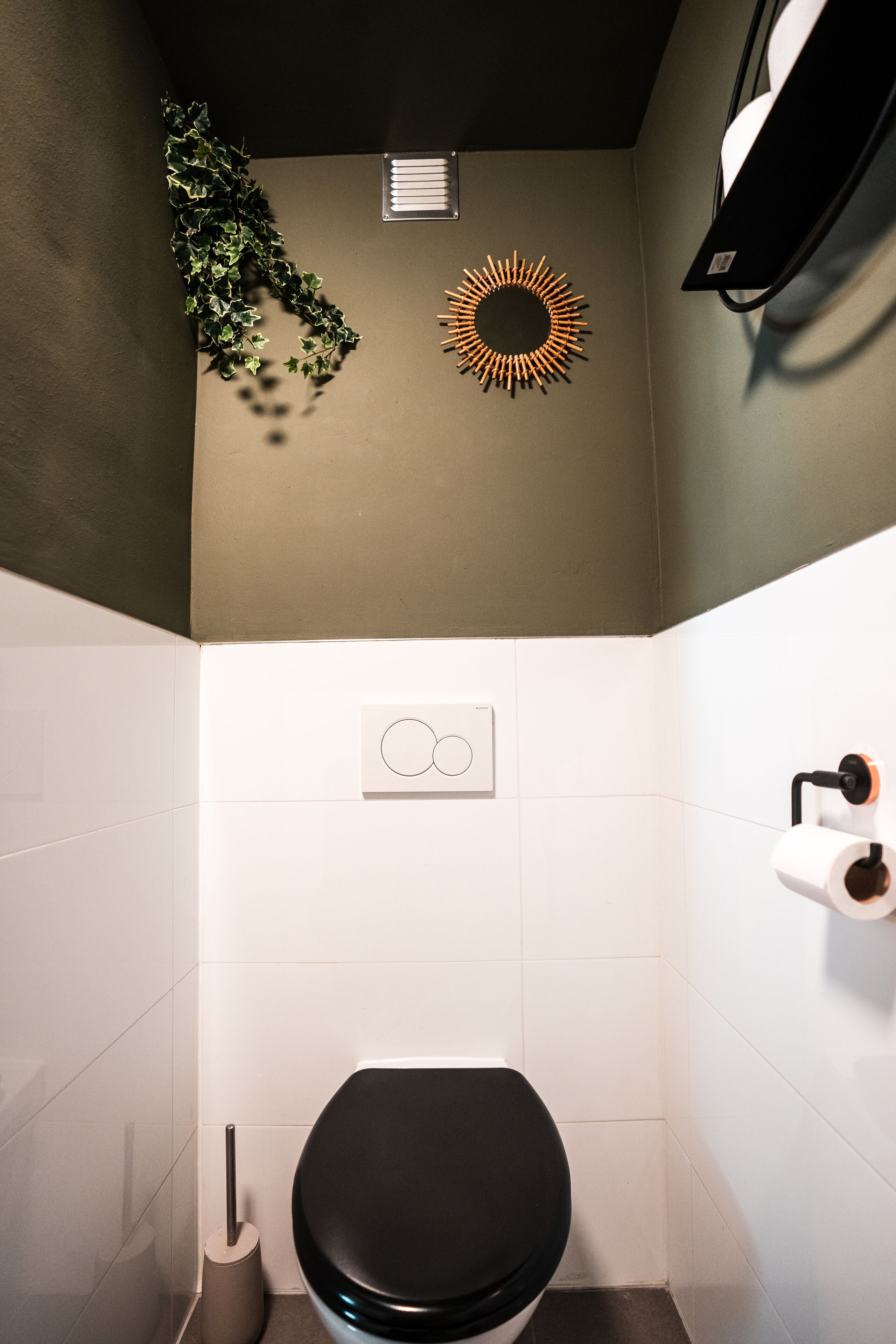 Ein Badezimmer mit Toilette, einem an der Wand befestigten Toilettenpapierhalter, einer Wandleuchte und einem dekorativen Spiegel im Sonnenstrahl-Design.