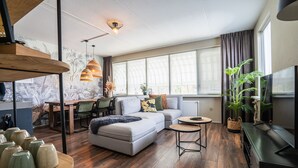 A modern living room with a sofa, coffee table, and dining area. There are plants, a TV, and a large window with curtains. - The Lion King (Groningen)