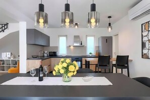 Eine moderne Küche mit Essbereich. Es gibt vier Pendelleuchten, einen Kühlschrank, einen Herd und eine Spüle. Der Esstisch ist mit einer Vase mit gelben Blumen und Weingläsern dekoriert.