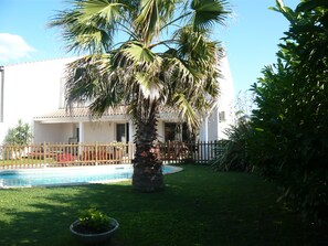 A pool surrounded by a fenced yard with a palm tree and a building with a tiled roof.