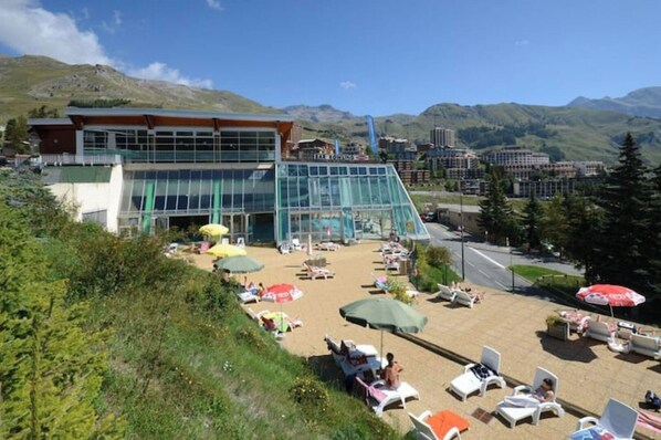 Un hôtel doté d’une façade en verre, d’un espace extérieur aménagé avec des parasols et surplombant un paysage montagneux.