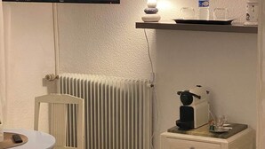 Una habitación pequeña con un radiador blanco, una mesa blanca con una silla, una mesita de noche blanca con una lámpara y un televisor montado en la pared.