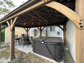A covered patio with a hot tub, outdoor seating, and a wooden pergola structure. - NEW! Updated *|* Stylish *|* OP getaway w/ Hot Tub (Overland Park)