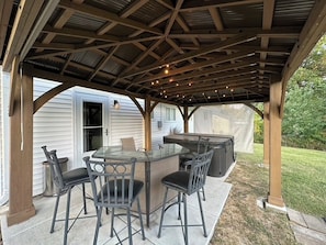A covered outdoor patio with a glass-top table and chairs, a grill, and a wooden pergola structure.
