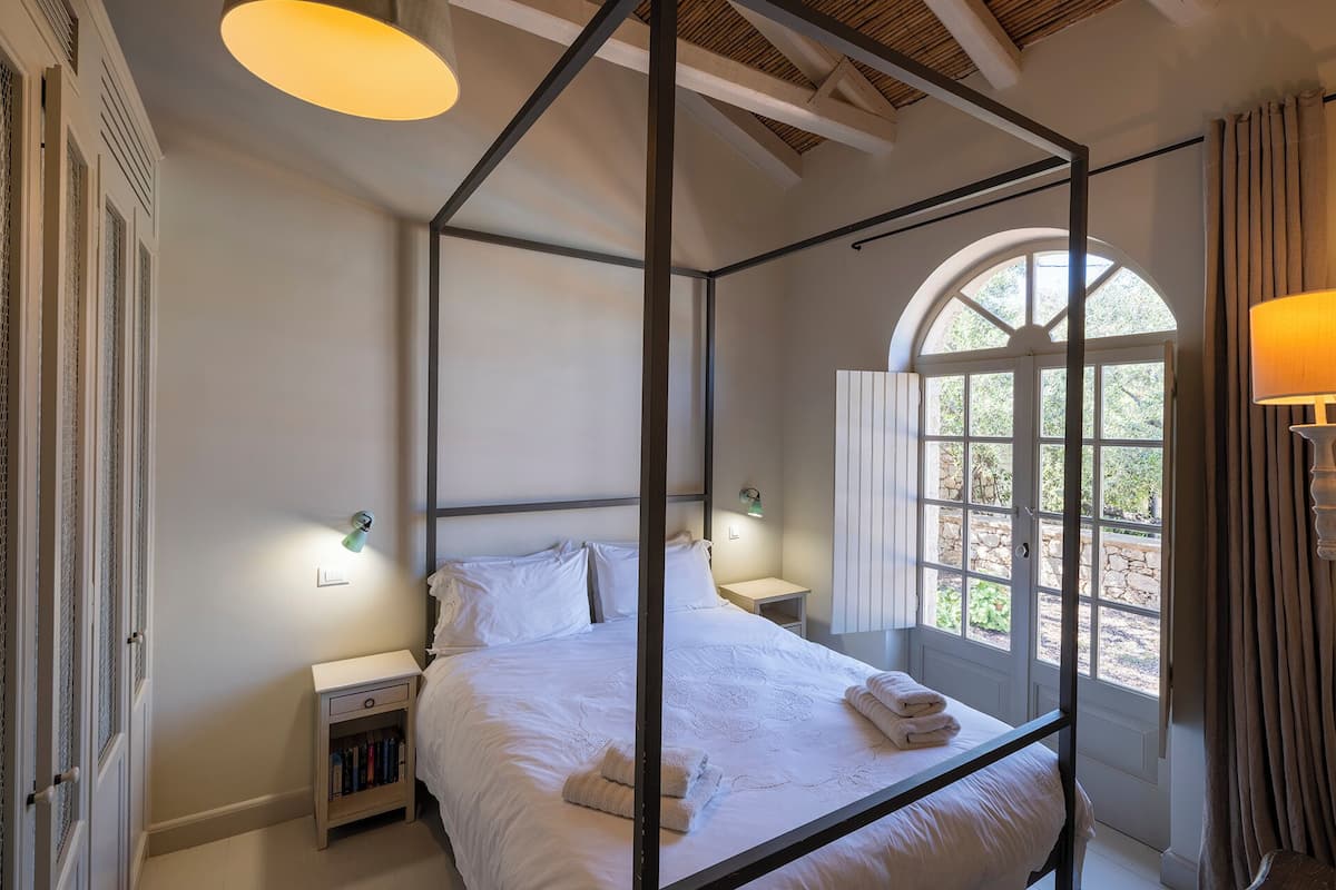 A four-poster bed with white linens and towels, a bedside table with a lamp, a nightstand with books, and a window with curtains.