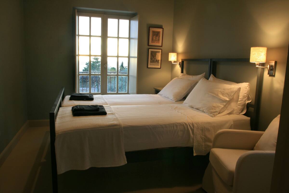 A hotel room with two beds, a chair, a window with a view, and wall-mounted lamps.