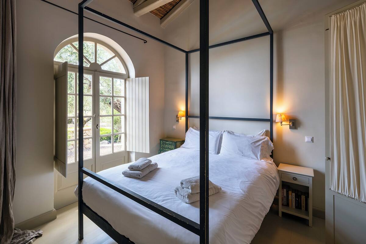 A bedroom with a four-poster bed, a bedside table with a lamp, and a window with curtains.