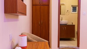 A hotel room with a wooden table, a glass-top side table, a TV mounted on the wall, and a wooden wardrobe.