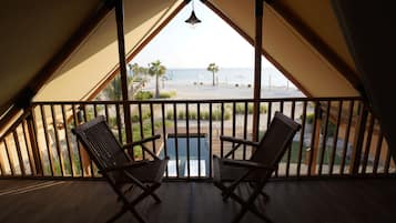 Un balcon avec des chaises et une table en bois, donnant sur une plage bordée de palmiers et offrant une vue sur un ciel dégagé.