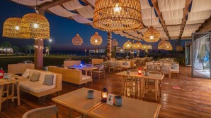 Un restaurant avec des tables et des chaises en bois, des lampes en osier suspendues et une vue sur la mer.