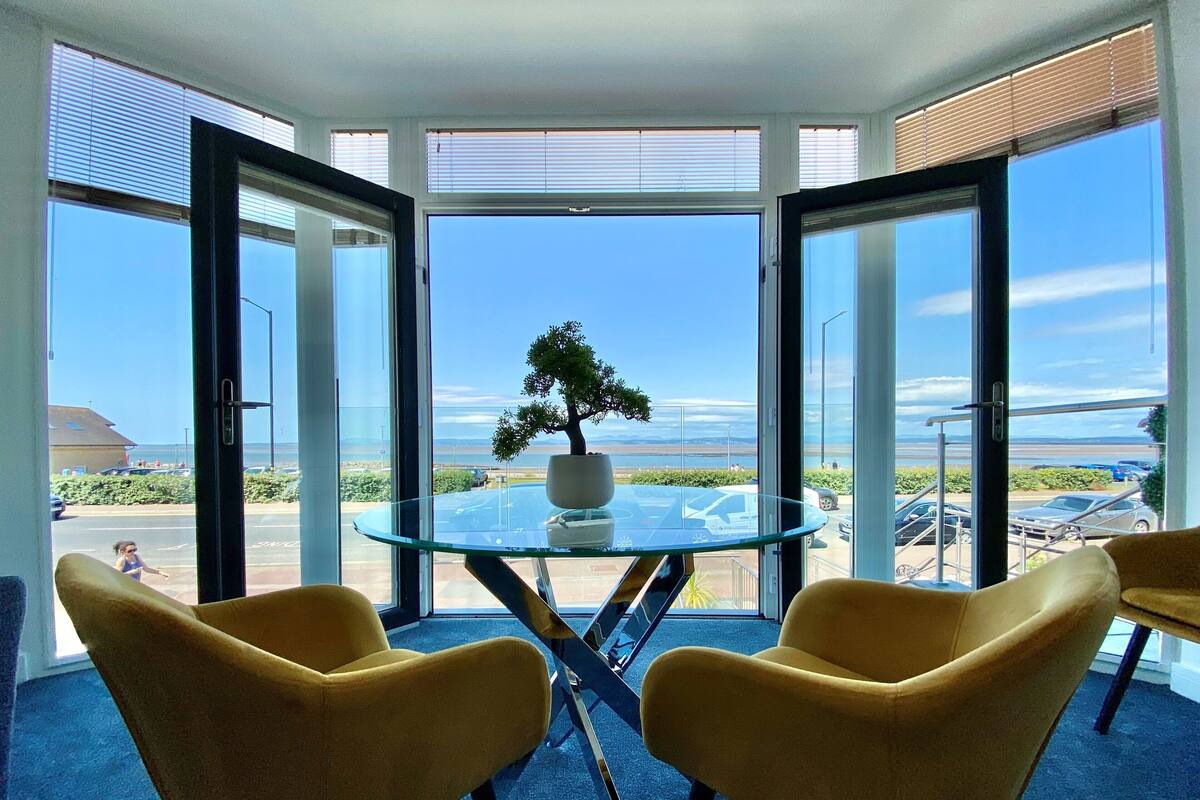 Ein Zimmer mit einem Glastisch, zwei gelben Stühlen, einer Bonsai-Pflanze und Blick auf Strand und Autos durch große Fenster.