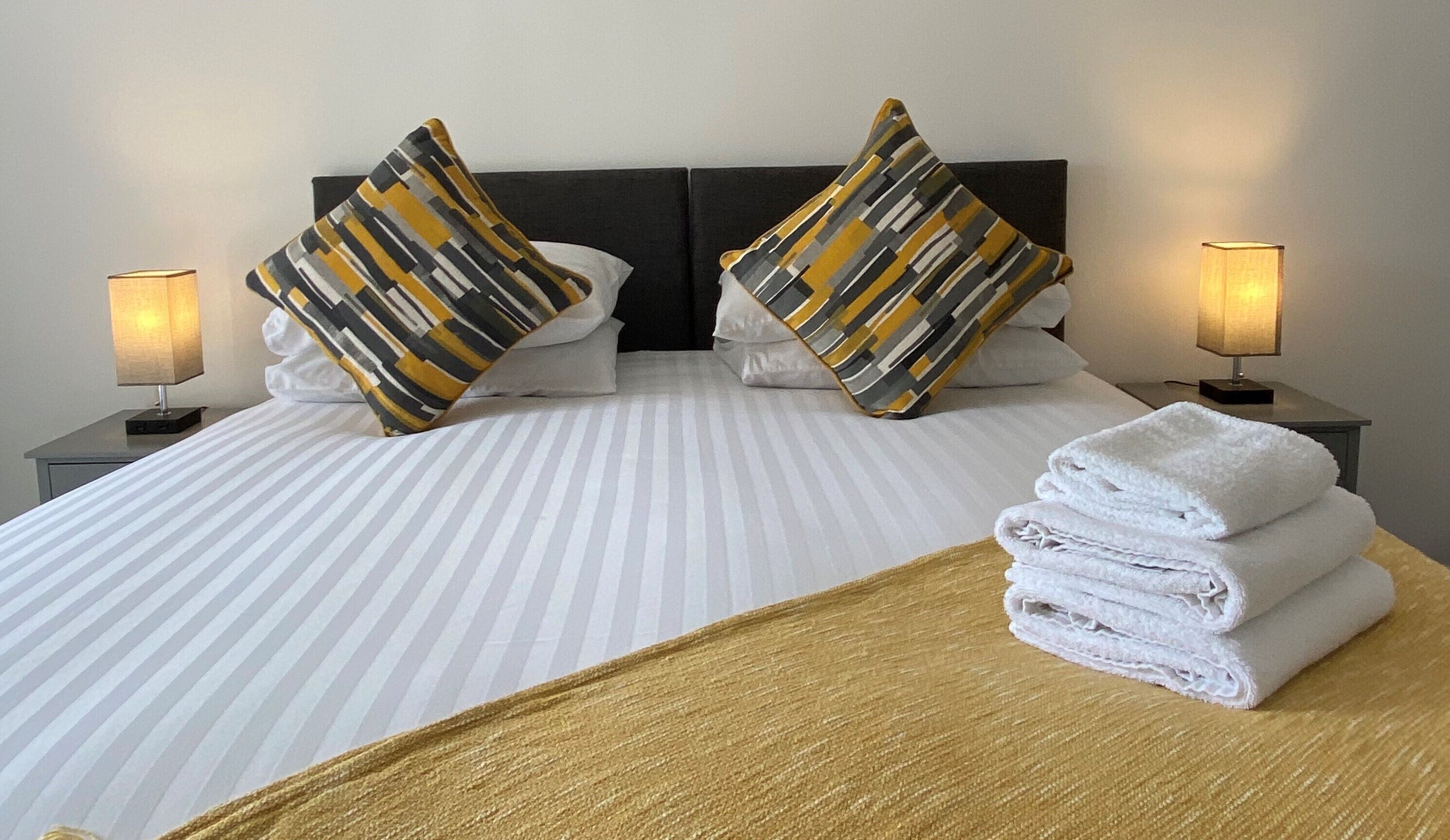 A hotel room with a bed, two pillows, two bedside lamps, and a stack of towels.