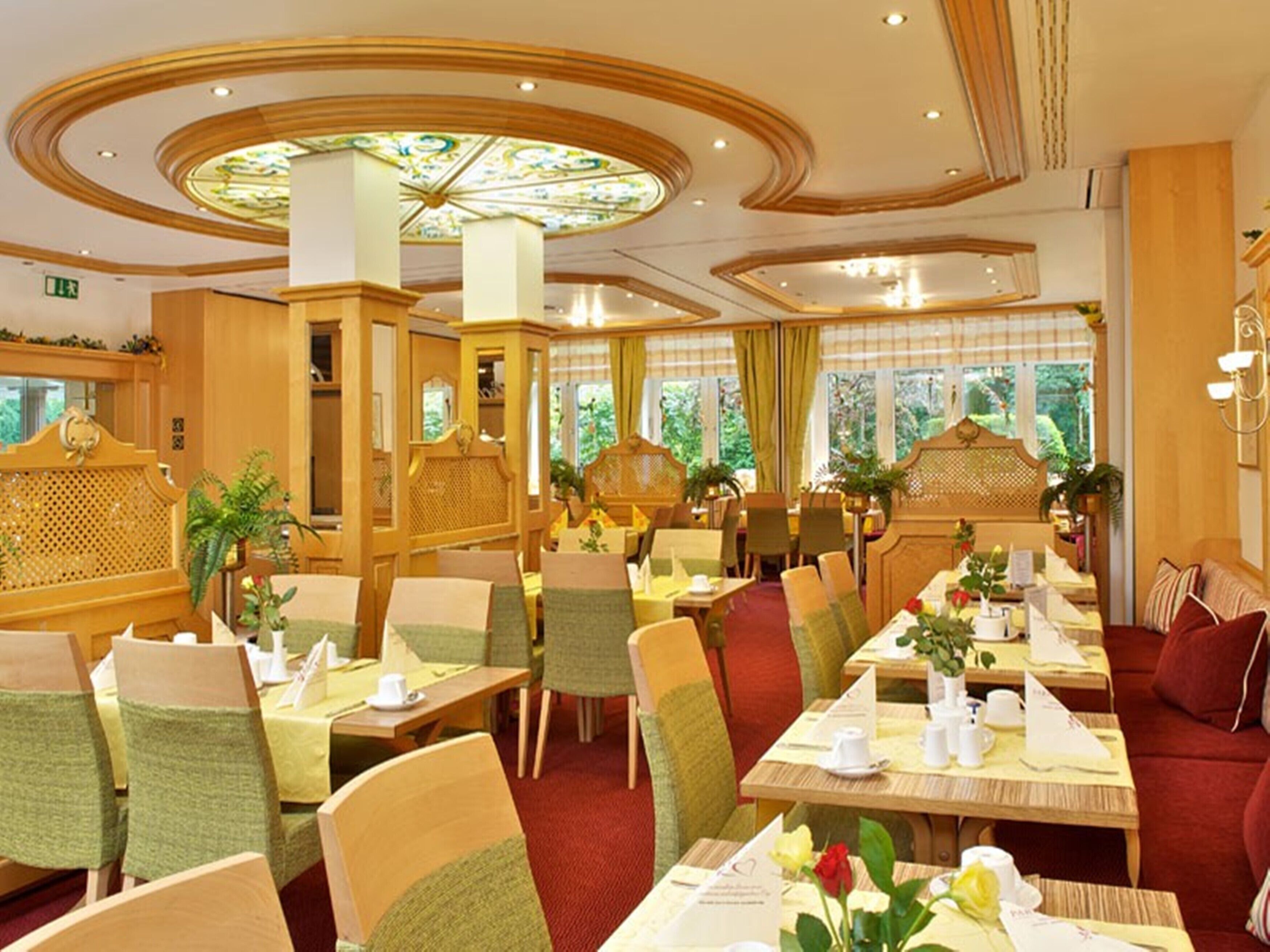 Un restaurante de diseño tradicional, con mobiliario de madera y una lámpara de araña en el centro. Las mesas están dispuestas con manteles blancos y centros de mesa florales.