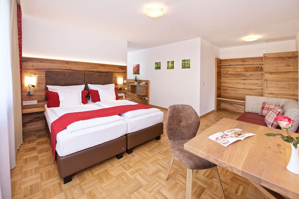 A modern hotel room with a large bed, a wooden headboard, a wooden desk, and a seating area with a sofa and armchair.