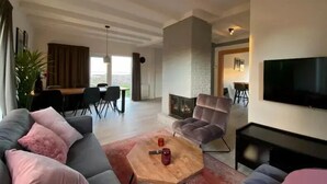 Een moderne woonkamer met een grijze bank, een houten salontafel en een flatscreen-televisie. Op de achtergrond is een eethoek met een tafel en stoelen.