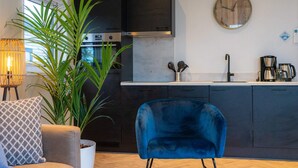 Een moderne keuken met donkere kastjes, een roestvrijstalen afzuigkap, een spoelbak en een koffiezetapparaat. Voorop staat een blauwe stoel en links staat een plant in een pot.