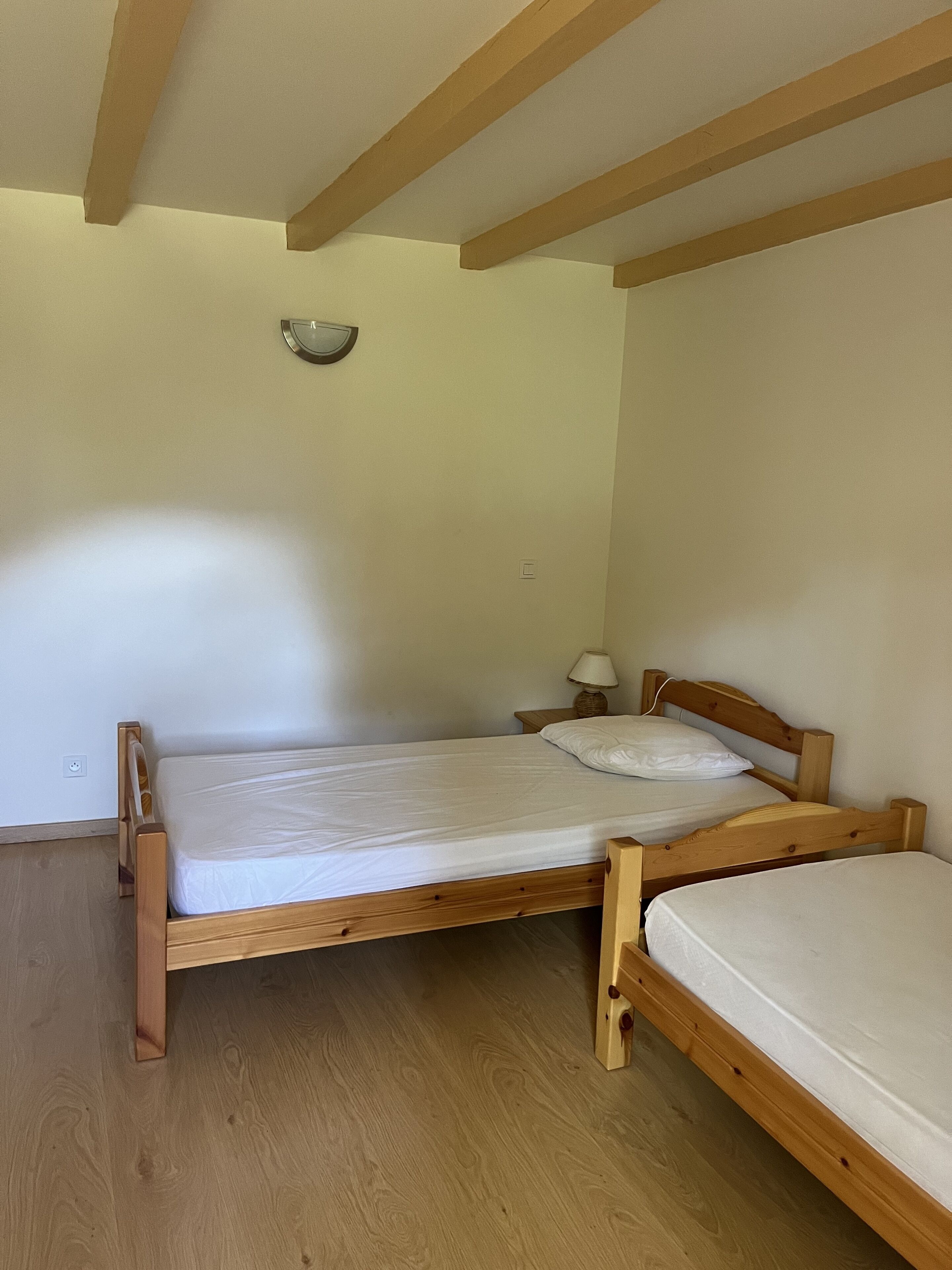 A small, single-room accommodation with two wooden beds, a nightstand with a lamp, and a wall-mounted light fixture.
