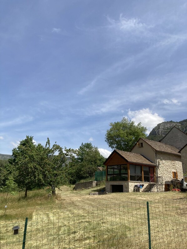 Une maison de campagne en pierre, dotée d’un porche en bois, entourée d’un champ clôturé et d’arbres.