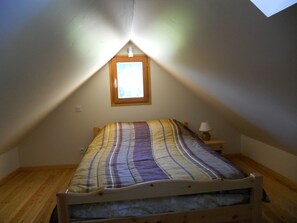 Un lit simple avec une couvre-lit à rayures, dans une pièce au plancher en bois, avec une table de chevet, une lampe et une fenêtre donnant sur des arbres.