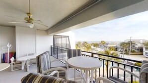 Un balcón con ventilador de techo, mobiliario blanco y vistas a la playa y edificios.