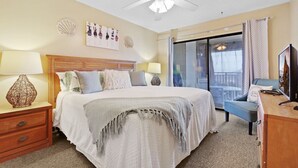 Habitación de hotel con una cama grande, dos mesitas de noche con lámparas, un ventilador de techo, un balcón con vistas y un televisor en un aparador de madera.