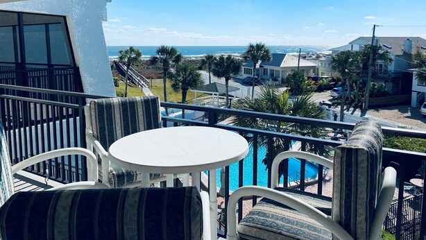 Un balcón con piscina, sillas blancas y vista al océano y la playa.