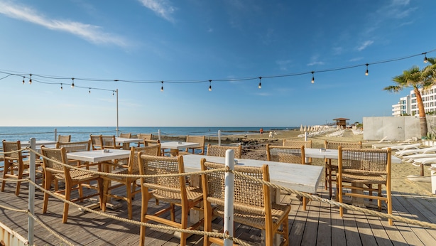 Un restaurante frente a la playa con terraza, mesas y sillas, con vista al océano.