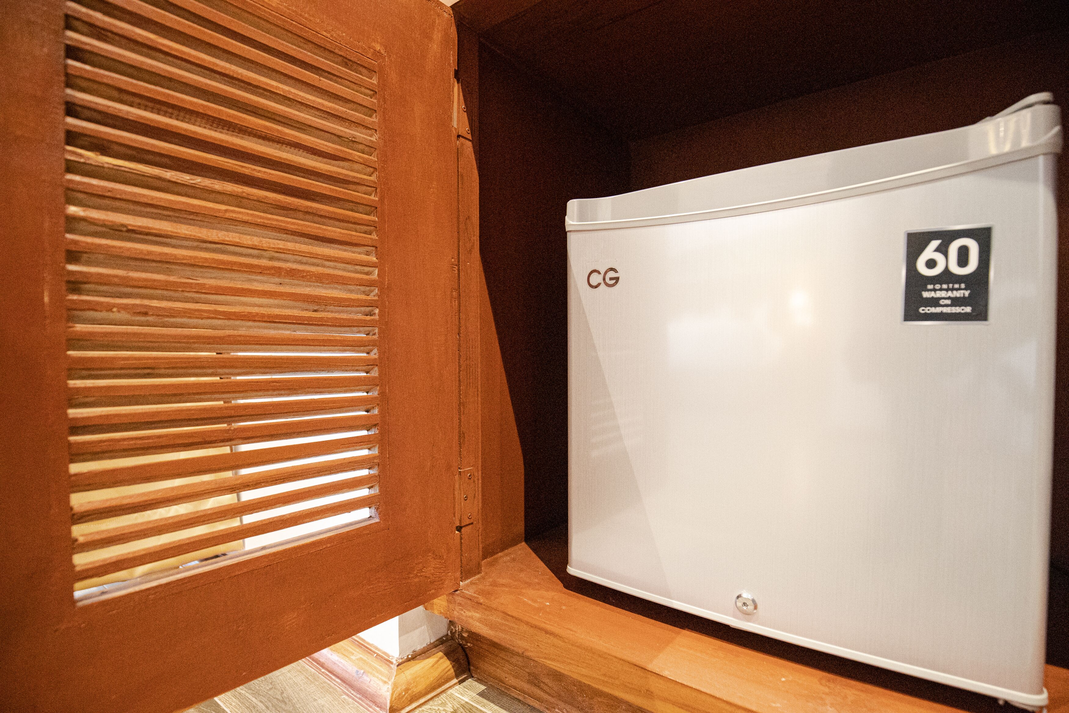 Un refrigerador blanco compacto con un rótulo que dice '60 meses de garantía', ubicado en un hueco con marco de madera y ventana con listones.